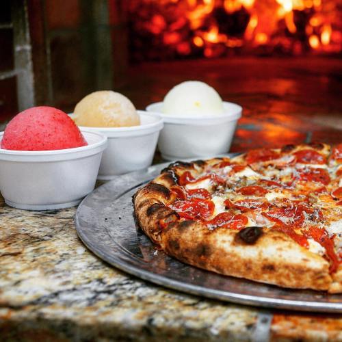 A pizza on a metal tray next to three bowls of colorful ice cream, with a warm, glowing fireplace in the background.