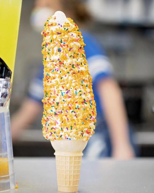 A tall ice cream cone covered in colorful sprinkles, set against a blurred background of an ice cream shop.