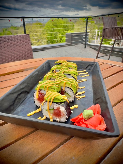 A platter of sushi rolls topped with avocado and drizzled with sauce, served with wasabi and pickled ginger on a wooden table.