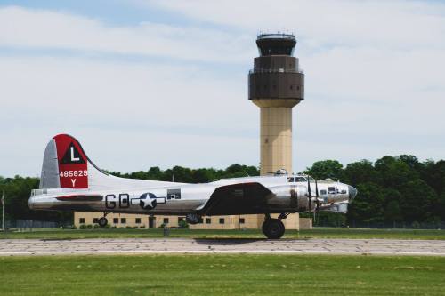 A vintage silver airplane with a red tail taxiing near a control tower on a sunny day.