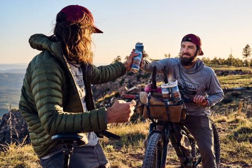 Two men on a mountain trail share drinks, one on a bike, enjoying a scenic view at sunset.