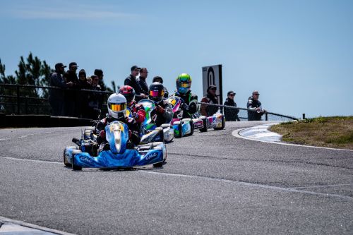 Four go-karts race on a track, with spectators watching from the sidelines under a clear blue sky.