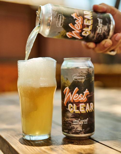 A hand pours a West Coast IPA from a can into a glass, creating a frothy head on the beer.