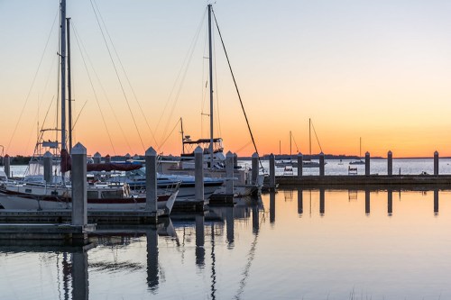 Sailboats docked at a marina during sunset, reflecting on calm waters with a colorful sky in the background.