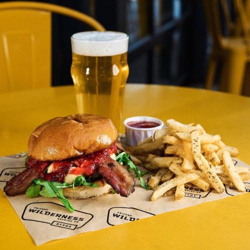 A delicious burger with bacon, lettuce, and sauce, served with fries and a pint of beer on a yellow table.