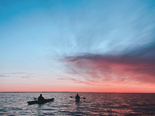 Two kayakers silhouette against a vibrant sunset over calm waters, with a gradient of blue and pink in the sky.