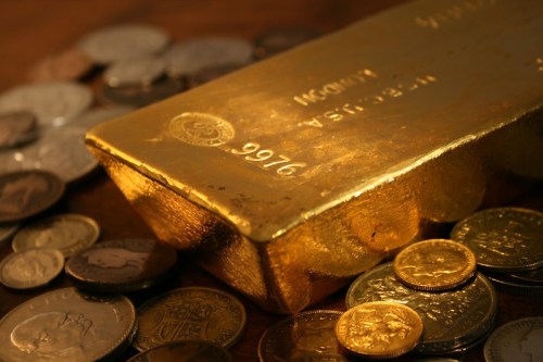 A gold bar surrounded by various coins on a wooden surface.