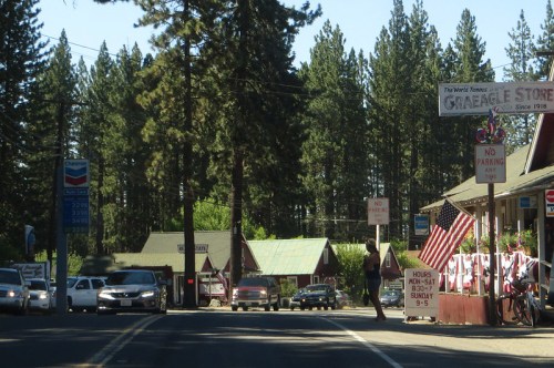 A small town street with a general store, vintage signs, and tall pine trees lining the road.