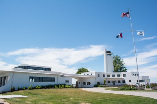 A modern white building with a tower, surrounded by green grass and flags, under a clear blue sky.