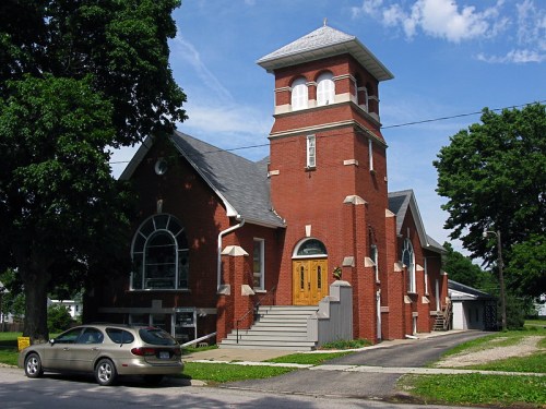 A red brick church with a tall tower, large windows, and a car parked in front, set against a blue sky and green trees.