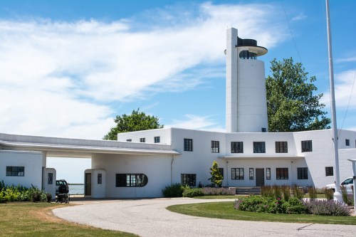 A modernist white building with a tall tower, surrounded by greenery and a clear blue sky.