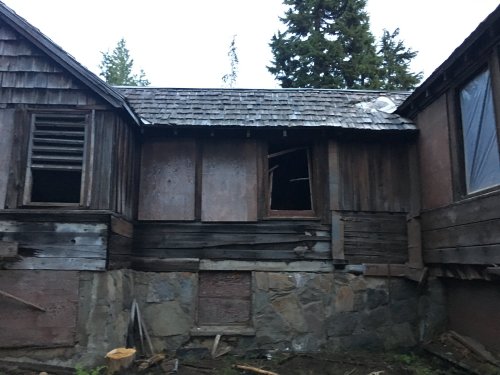 Abandoned wooden house with broken windows and weathered walls, surrounded by trees in a dimly lit setting.