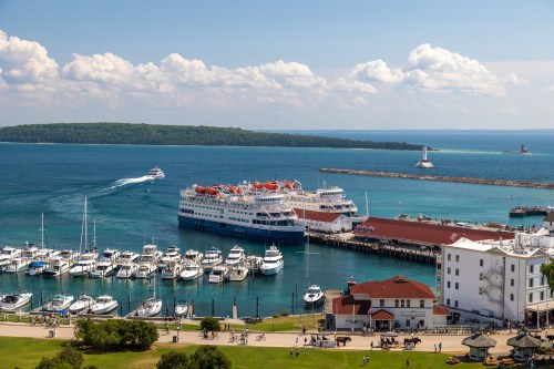 A scenic harbor with a large cruise ship, smaller boats, and a lighthouse under a blue sky with fluffy clouds.