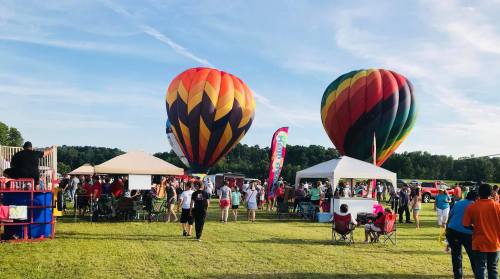 Colorful hot air balloons rise above a crowd at a festival, with tents and people enjoying the outdoor event.