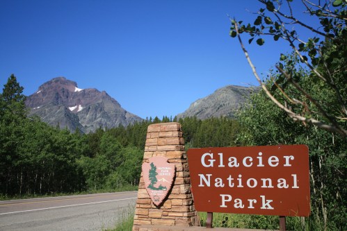 Sign for Glacier National Park with mountains and clear blue sky in the background. Lush greenery surrounds the area.