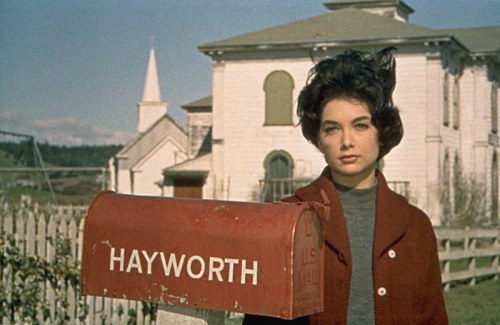 A woman stands by a red mailbox labeled "HAYWORTH" in front of a white building and a church in the background.