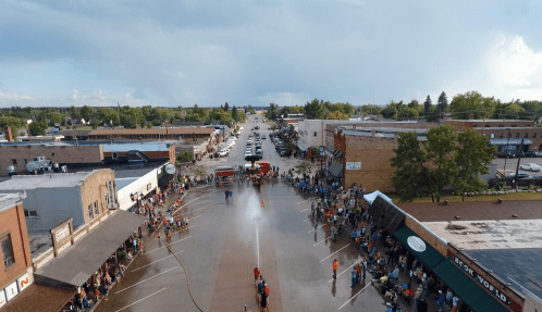 Aerial view of a bustling street festival with crowds, buildings, and a fire truck spraying water in the center.