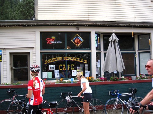 A quaint café with cyclists outside, featuring a sign that reads "Jamestown Mercantile Co. Café" and colorful flowers.
