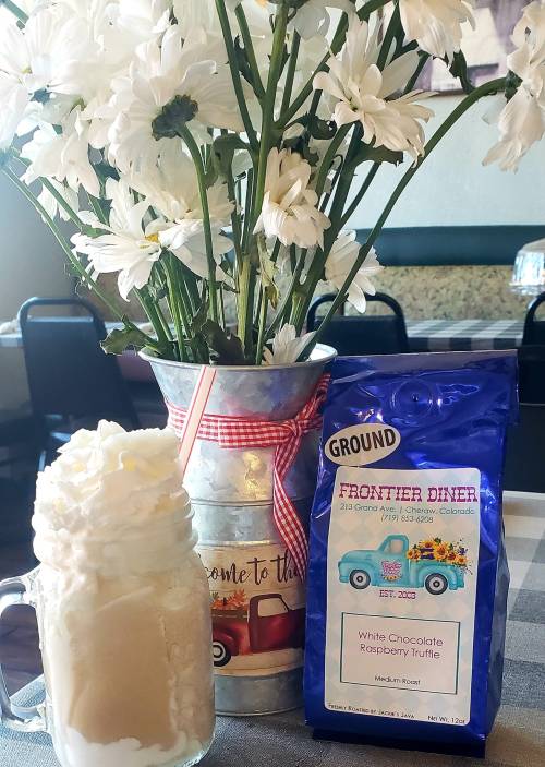 A mason jar filled with a creamy drink topped with whipped cream, next to a vase of flowers and a coffee bag.