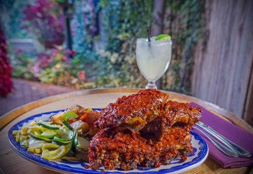 A plate of barbecue ribs with vegetables and a cocktail in a vibrant, outdoor setting.