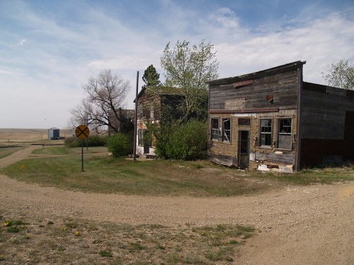 Abandoned wooden buildings at a rural intersection, with a dirt road and a railroad crossing sign nearby.