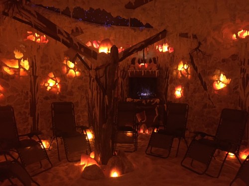 A cozy room with glowing salt lamps, sandy floor, and chairs arranged around a fireplace, creating a warm ambiance.