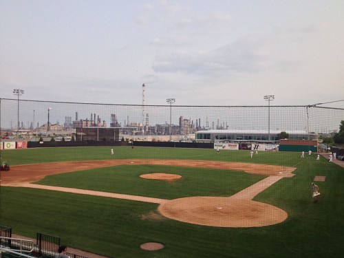 A baseball field with a grassy infield and dirt diamond, set against an industrial skyline in the background.
