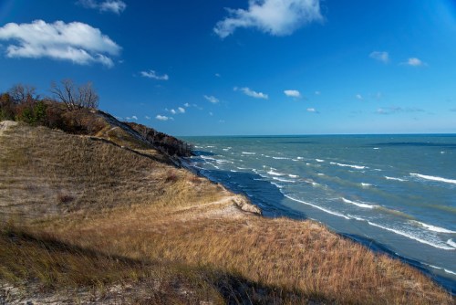 A scenic view of a grassy shoreline meeting a wavy blue lake under a clear sky with scattered clouds.