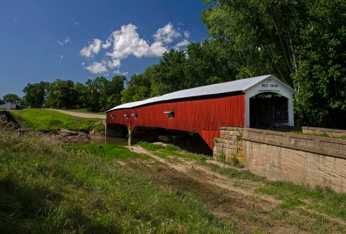 A red covered bridge spans a small creek, surrounded by green trees and a clear blue sky with fluffy clouds.