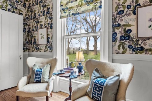 Cozy room with floral wallpaper, two armchairs, a side table, and a window view of trees and a fence outside.