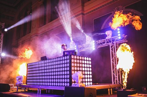 A DJ performs on stage with a light display and flames, creating an energetic atmosphere at a live event.