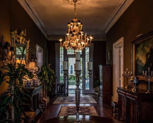 A dimly lit hallway with a chandelier, elegant decor, and large windows showcasing greenery outside.