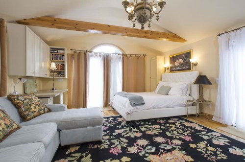 Cozy bedroom with a bed, armchair, floral rug, and large windows, featuring warm lighting and elegant decor.