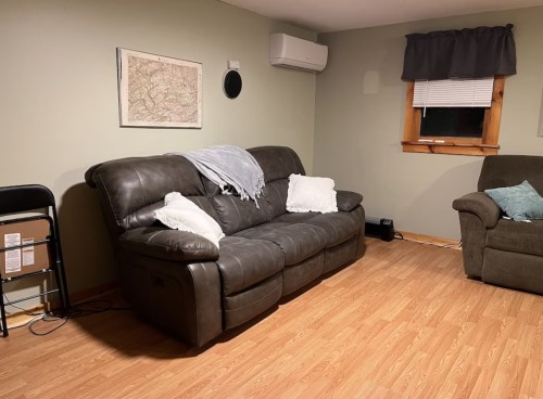 A cozy living room with a dark leather couch, a chair, and a wall-mounted air conditioner. A map hangs on the wall.