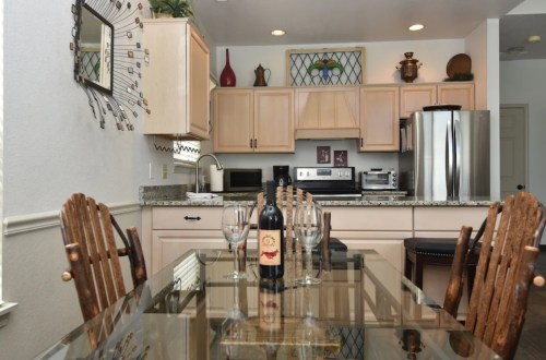 A modern kitchen with wooden chairs, a glass dining table, wine glasses, and a bottle of wine on the table.
