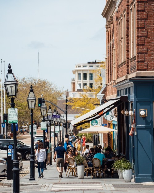 A bustling street scene with people walking, dining outdoors, and historic buildings under a clear sky.