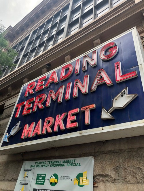 Sign for Reading Terminal Market with bright red letters and directional arrows, set against a stone building.