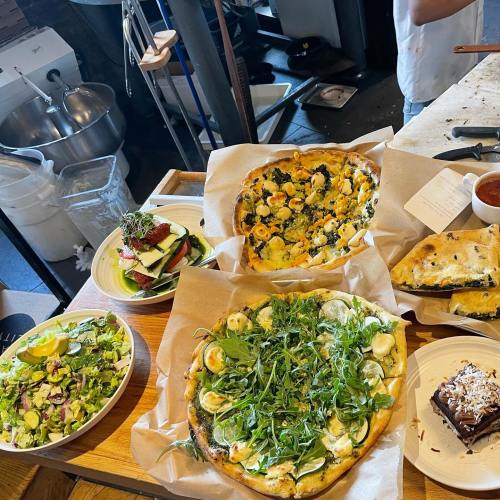 A table with two pizzas topped with greens, a salad, a layered vegetable dish, and a brownie dessert.