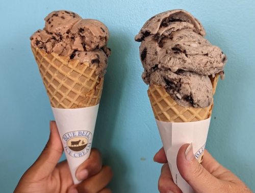 Two hands hold ice cream cones with chocolate and cookies and cream flavors against a blue background.