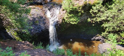 A small waterfall cascades into a dark pool, surrounded by lush greenery and rocky terrain.