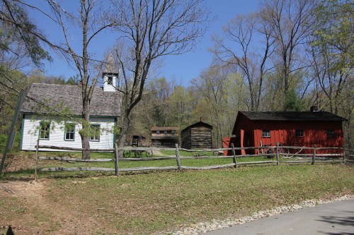 A rustic scene featuring a white building with a bell tower, a red barn, and a wooden cabin, surrounded by trees.