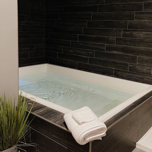 A modern spa with a large, inviting hot tub and neatly folded towels beside it, surrounded by dark tiled walls.