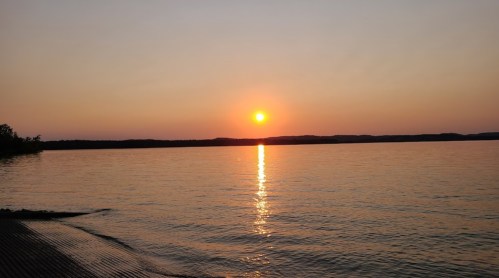 A serene sunset over a calm lake, with the sun reflecting on the water and distant hills in the background.