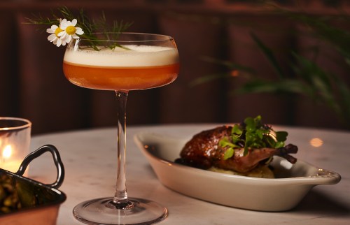 A cocktail garnished with a flower beside a plate of roasted chicken on a marble table. Soft lighting enhances the ambiance.