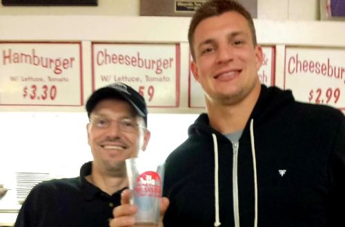 Two men smiling together in a diner, one holding a glass, with menu items visible in the background.