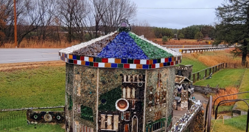 A colorful, mosaic-covered building with a unique roof design, surrounded by grass and trees along a road.