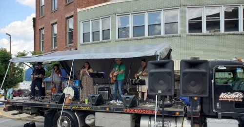 A band performs on a truck stage in front of a building, with musicians playing instruments and singing.