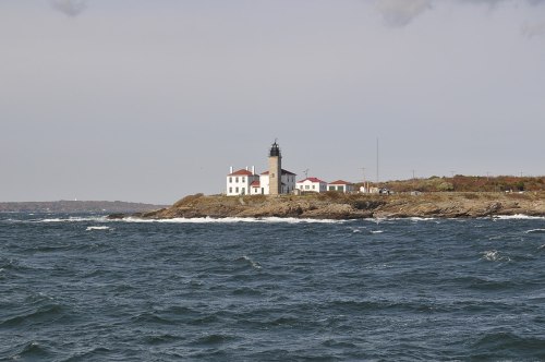 A lighthouse on a rocky shore, surrounded by waves and a cloudy sky, with buildings nearby.