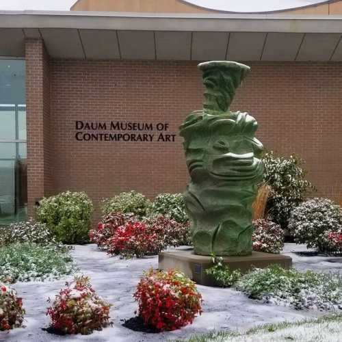 A green sculpture stands in front of the Daum Museum of Contemporary Art, surrounded by snow and colorful plants.