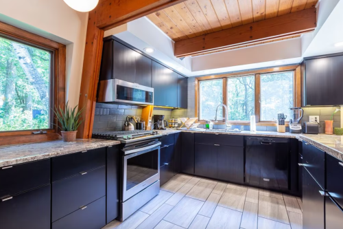 Modern kitchen with dark cabinets, granite countertops, and large windows overlooking a forested area.